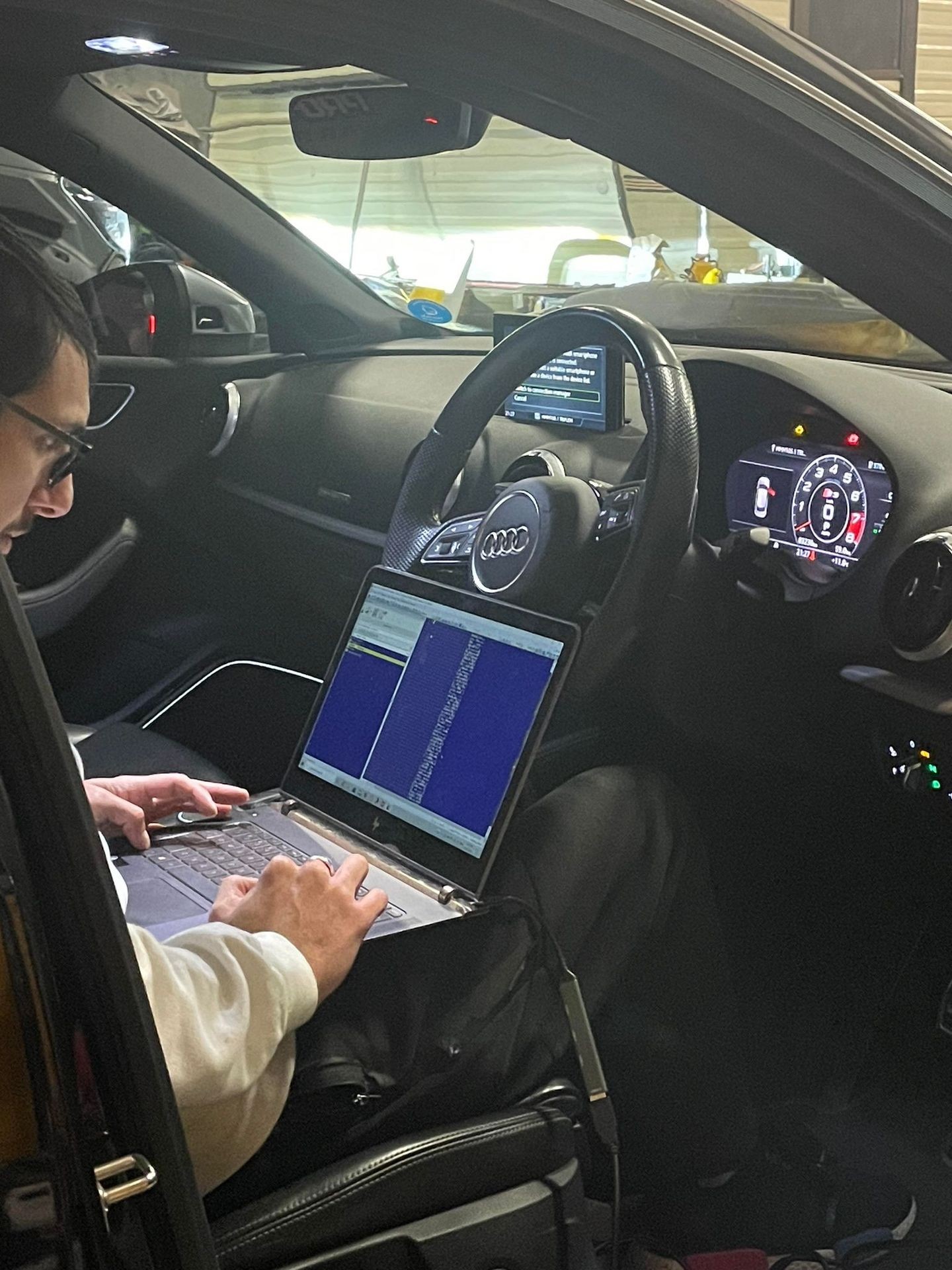 Person using a laptop in the driver's seat of a car with dashboard displays visible.